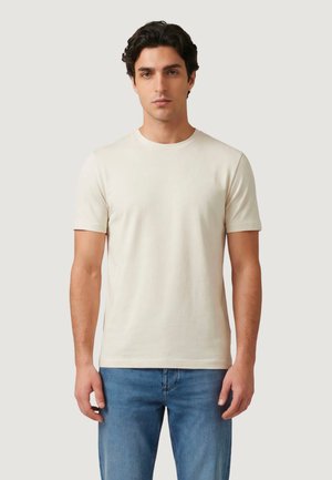 Giovane uomo con capelli scuri che indossa una maglietta beige chiaro a maniche corte e jeans blu, in piedi davanti a uno sfondo neutro.