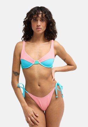 Kobieta z kręconymi ciemnymi włosami, ubrana w kolorowy bikini w różowym, niebieskim i zielonym kolorze, stojąca z jedną ręką na biodrze na jednolitym tle.