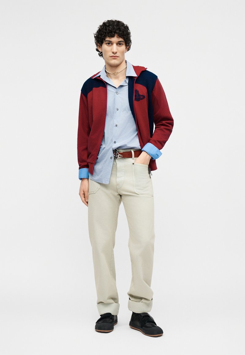 Young man with curly hair wearing red and navy jacket, light blue striped button-up shirt, beige pants, and black shoes, standing with hand in pocket.