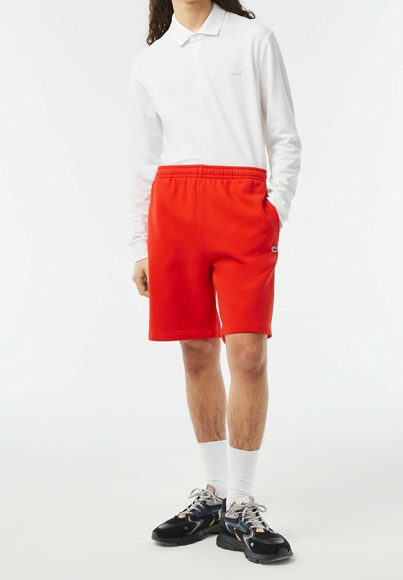 Personne portant un short rouge vif, un polo blanc à manches longues, des chaussettes blanches et des chaussures de sport noires, debout devant un fond neutre.
