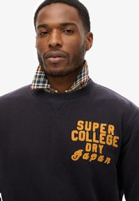 Sudadera de color marino con texto bordado "SUPER COLLEGE DRY Japan", tejido texturizado, con un cuello de cuadros en rojo, verde y azul.