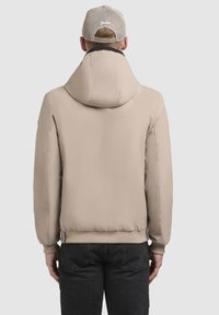 Giacca bomber beige con cappuccio, caratterizzata da una texture liscia, polsini elasticizzati e una leggera lucentezza, abbinata a jeans scuri.