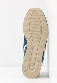 Semelle en caoutchouc beige avec motifs de grip texturés et logo New Balance sur la semelle bleue et blanche d’une chaussure de sport.