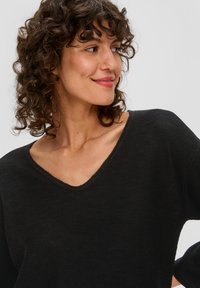 Schwarzer Strickpullover mit V-Ausschnitt, lockerer Passform und Drei-Viertel-Ärmeln. Die Textur wirkt weich und leicht gerippt.