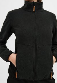 Czarna kurtka zapinana na zamek z teksturowanej tkaniny, z wysokim kołnierzem i dwiema bocznymi kieszeniami; wyposażona w drewniane uchwyty do zamka.