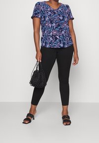 Blusa blu navy con motivo paisley rosa e bianco; design a scollo a V. Indossata con pantaloni neri aderenti e una borsa nera a rete testurizzata. Sandali neri con tacco.