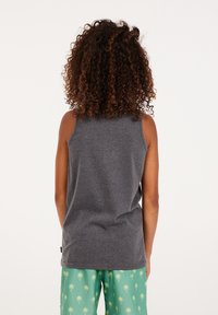 Bambino con capelli ricci indossa una maglietta senza maniche grigia scura e pantaloni corti verdi con motivi di palme, in piedi di fronte a uno sfondo bianco.