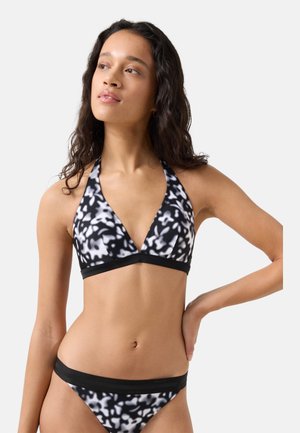 Frau mit langem, dunklem, lockigem Haar, die ein schwarz-weiß gemustertes Neckholder-Bikini-Oberteil und passende Unterteile trägt und mit einer Hand an der Hüfte posiert.