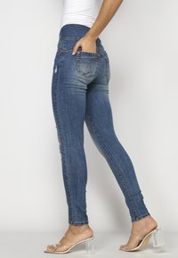 Femme portant un jean skinny bleu délavé et des sandales à talons hauts transparentes, une main dans la poche arrière, debout devant un fond uni.