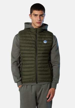 Giovane uomo che indossa un gilet trapuntato verde oliva sopra un felpa grigia con cappuccio e pantaloni della tuta abbinati, in piedi con le mani in tasca su uno sfondo bianco.