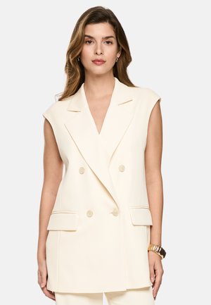 Junge Frau trägt einen ärmellosen, doppelt geknöpften cremefarbenen Blazer mit Klappentaschen und einem goldenen Armband am rechten Handgelenk.