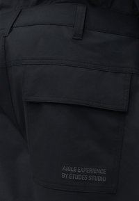 Gros plan sur un pantalon en tissu noir avec une grande patte de poche et un texte brodé "AIGLE EXPERIENCE BY ÉTUDES STUDIO" sur la poche.
