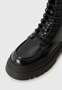 Bottine en cuir noir avec un bout arrondi, semelle épaisse et un système de laçage prononcé. Finition lisse avec des accents texturés sur la semelle.