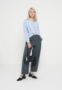 Chemise boutonnée bleu clair avec une texture subtile, pantalon gris à jambes larges, sac à main noir et chaussures noires à bout pointu.