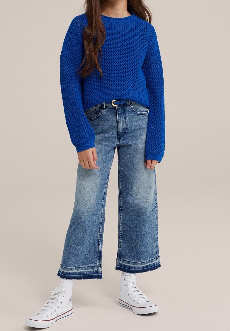 Enfant portant un pull en maille bleu vif, un jean large délavé avec des ourlets bruts, des chaussettes blanches et des baskets montantes blanches, debout sur un fond neutre.