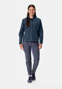 Marinblå vattentät jacka med dragkedja, elastiska muddarna; kombinerad med ljusgrå byxor med fickor och sidodetaljer. Svarta skor.