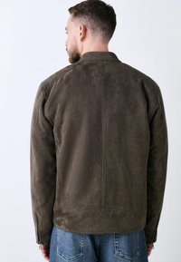 Chaqueta de ante marrón con una textura suave, con puños con cremallera y un cuello sencillo, combinada con jeans de mezclilla azules.