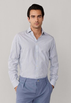 Man die een blauw-wit gestreept shirt met lange mouwen draagt, ingestopt in lichtblauwe op maat gemaakte broek, staand met handen in de zakken.