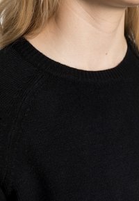 Black knit sweater featuring a ribbed crew neckline and soft texture, shown from the side against light skin tone.