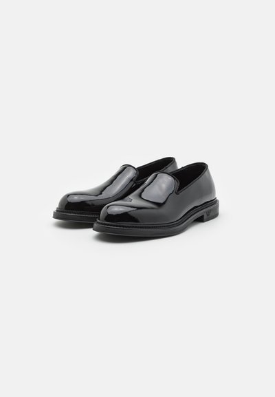 Schwarze Lackleder-Slipper mit runder Zehenform, glatter Oberfläche, elastischen Seiten und einer subtilen strukturierten Sohle. Keine sichtbaren Logos.