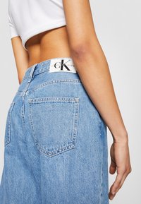 Person som bär ljusblå Calvin Klein-jeans och en vit crop top, vänd med ryggen och armen på sidan mot en neutral bakgrund.