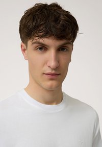 Witte katoenen T-shirt met ronde hals, gladde textuur en een klein logo op de linkerkant van de borst. Model toont het shirt tegen een neutrale achtergrond.
