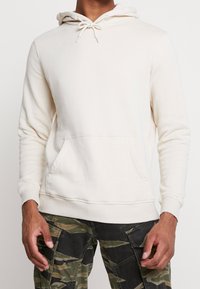 Krémový pulover s kapucí vyrobený z měkké tkaniny, s nastavitelnou kapucí, přední klokankovou kapsou a žebrovanými manžetami a lemem.