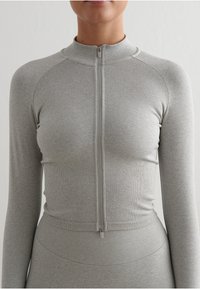 Sudadera de manga larga con cremallera en gris claro, que presenta un diseño ajustado, una sutil textura acanalada y un cuello alto. Ideal para ropa deportiva.