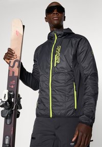 Melnā siltinātā slēpošanas jaka ar zaļiem rāvējslēdzēju akcentiem, ar kviltotu tekstūru, divām sānu kabatām un kapuci. Modelis tur slēpes.