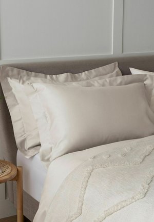 Beige Kissen und passende Bettdecke auf einem grau gepolsterten Bett mit einem hölzernen Beistelltisch in einem sanft beleuchteten Schlafzimmer-Eck.