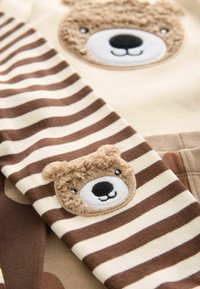 Ensemble en coton comprenant un haut beige avec une pièce en patch représentant un visage d'ours duveteux et des manches rayées marron et crème. Texture douce et design ludique.