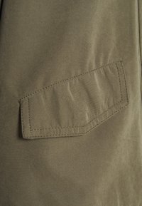 Olive green fabric with a smooth texture, featuring a square flap pocket stitched with contrasting thread in a diagonal position.