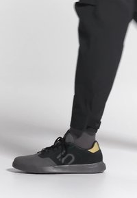 Henkilö, jolla on mustat ja harmaat lenkkitossut keltaisilla yksityiskohdilla, tummanharmaat sukat ja mustat housut, seisomassa valkoisella pinnalla.