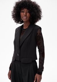 Czarna krótką marynarkę z jednym guzikiem, z dwiema przednimi kieszeniami, nałożoną na przezroczystą bluzkę z długim rękawem z kwiecistym haftem.