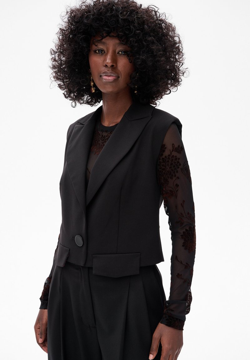 Czarna krótką marynarkę z jednym guzikiem, z dwiema przednimi kieszeniami, nałożoną na przezroczystą bluzkę z długim rękawem z kwiecistym haftem.