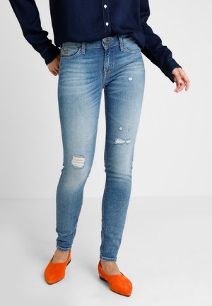 Žena v modrých světlých roztrhaných džínách s běžným pasem, tmavě modrém topu s dlouhým rukávem a jasně oranžových balerínách stojí před bílým pozadím.