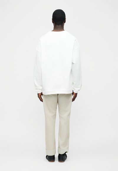 Άνδρας που στέκεται με την πλάτη στραμμένη, φοράει ένα απλό λευκό πουκάμισο με μακριά μανίκια, μπεζ παντελόνια με μανσέτες και μαύρα παπούτσια πάνω σε λευκό φόντο.