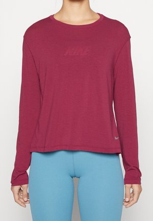 Femme portant un t-shirt Nike à manches longues bordeaux et des leggings bleu clair, debout devant un fond blanc uni.