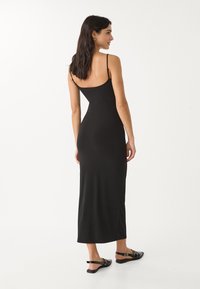 Robe maxi noire ajustée avec des fines bretelles et un dos plongeant. Texture lisse, ourlet à hauteur des genoux, portée avec des sandales plates noires.