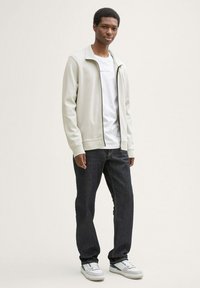 Veste zippée grise claire sur un t-shirt blanc, assortie à un jean foncé et des baskets blanches. Design moderne avec des caractéristiques minimalistes.