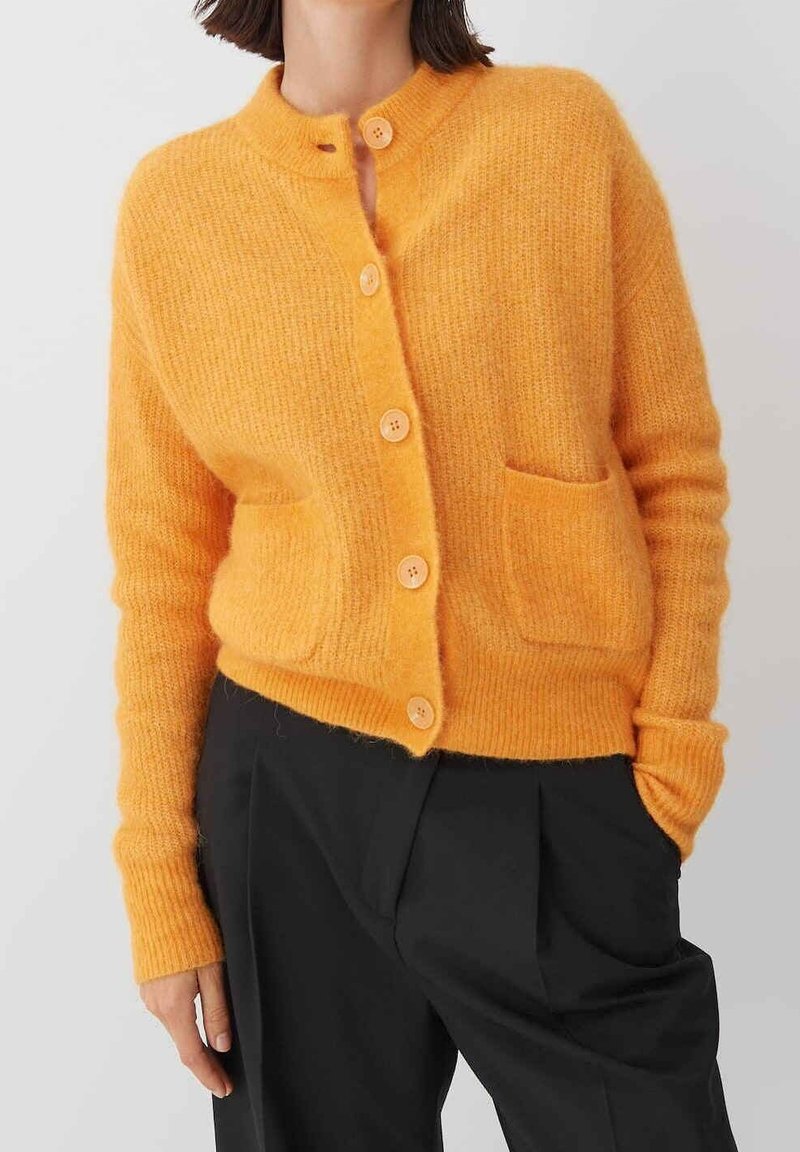 Orange, texturerad cardigan med rund krage, fem knappar och en framficka. Bärs över svarta byxor, vilket framhäver dess varma färg.