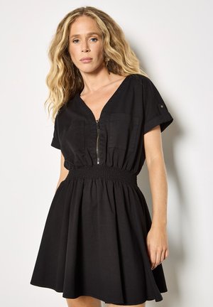 Femme aux longs cheveux blonds ondulés portant une robe noire à manches courtes avec une fermeture éclair à l'avant et une taille froncée, debout contre un mur blanc.