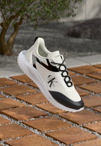 Hvit og svart sneaker med en strømlinjeformet design, mesh-overdel, rund tå og en fremtredende logo på siden. Teksturert såle med demping.