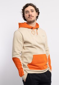 Sudadera beige con acentos naranjas en las mangas y el bolsillo. Tejido suave, capucha ajustable con cordón y detalle del logo en el pecho.