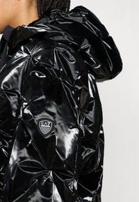 Μαύρο μπουφάν τύπου puffer με γυαλιστερό φινίρισμα, διαθέτει κουκούλα και σχεδιασμό σε κουτιά. Περιλαμβάνει ταμπέλα με το λογότυπο EA7 στο μανίκι.