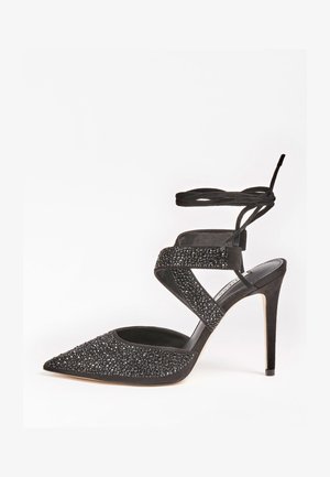 Escarpin noir à talons hauts avec un bout pointu, un corps en suède texturé, des embellissements en cristal et des brides croisées à la cheville.