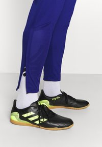 Czarne buty sportowe z gumowymi podeszwami, z akcentami neonowej żółci i trzema paskami. Noszone z purpurowymi spodniami i szarymi skarpetami.