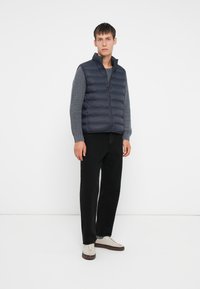 Junger Mann, der eine marineblaue Steppweste über einem grauen Strickpullover, schwarzen Jeans und weißen Turnschuhen trägt und vor einem weißen Hintergrund steht.