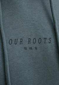 Mørkegrønn hettegenser laget av mykt stoff. Har "OUR ROOTS"-logo trykt i svart. Inkluderer teksturert snor til hetten.