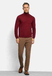 Burgunderfarbener Rollkragenspullover, kombiniert mit khakifarbener Hose und braunen Lederschuhen. Der Pullover hat eine figurbetonte Passform und eine gerippte Textur an den Bündchen und dem Saum.